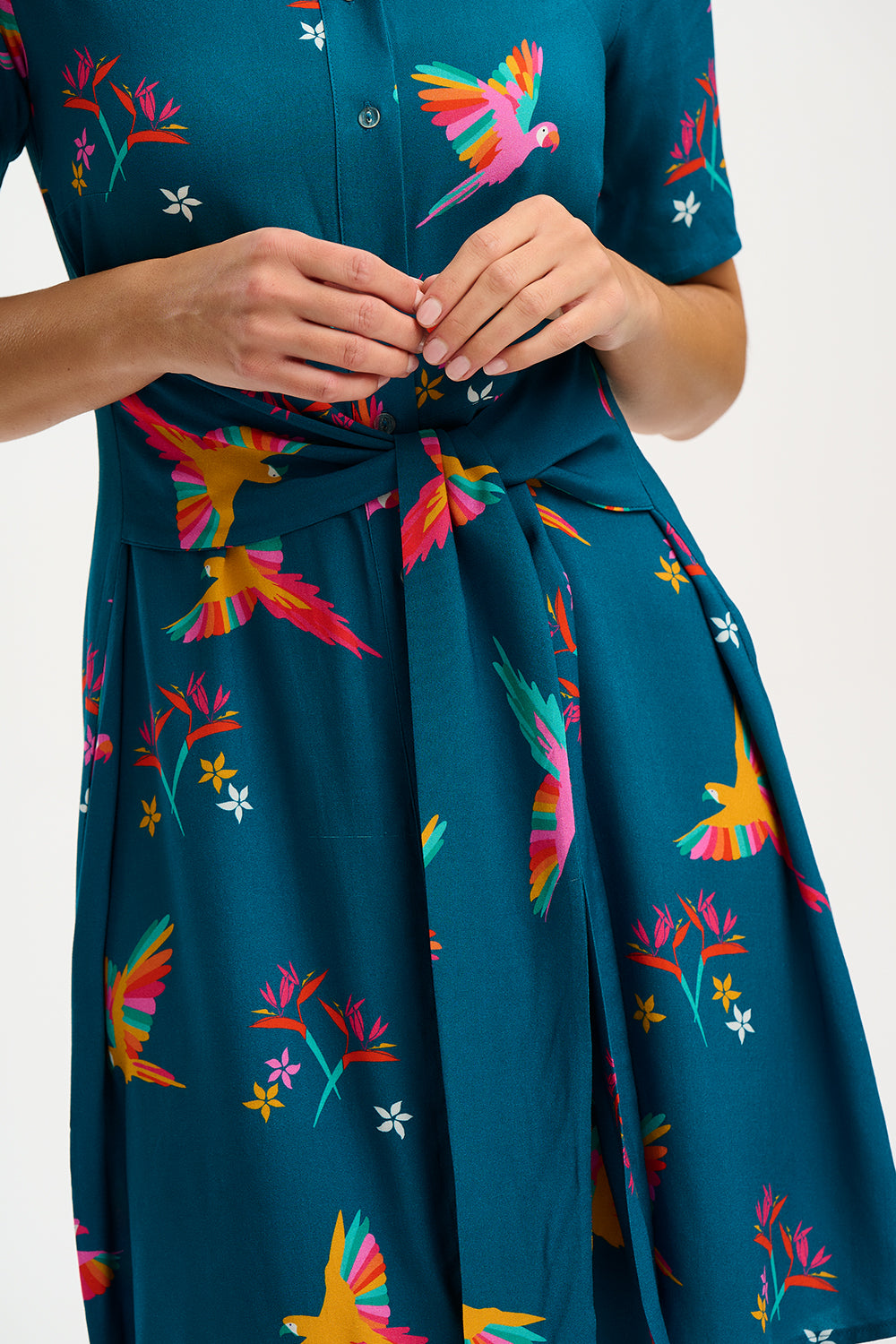 A model wearing a teal shirt dress with a Rainbow Parrots print, featuring short sleeves and a tie-front belt.