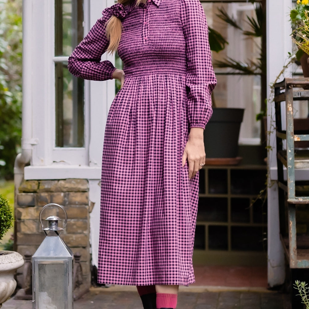 A woman stands on stone steps in front of a house doorway wearing a long-sleeved purple and white gingham midi dress with a smocked waist, paired with black rain boots and pink socks.