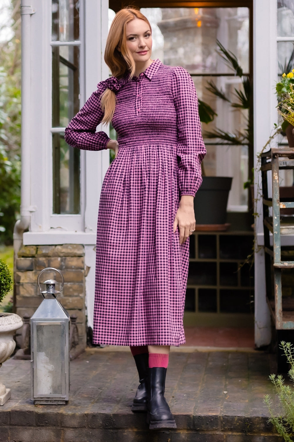 A woman stands on stone steps in front of a house doorway wearing a long-sleeved purple and white gingham midi dress with a smocked waist, paired with black rain boots and pink socks.