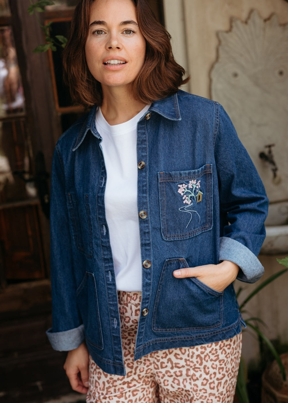 Woman wearing a blue denim jacket with floral pocket detail, white shirt, and patterned pants indoors.