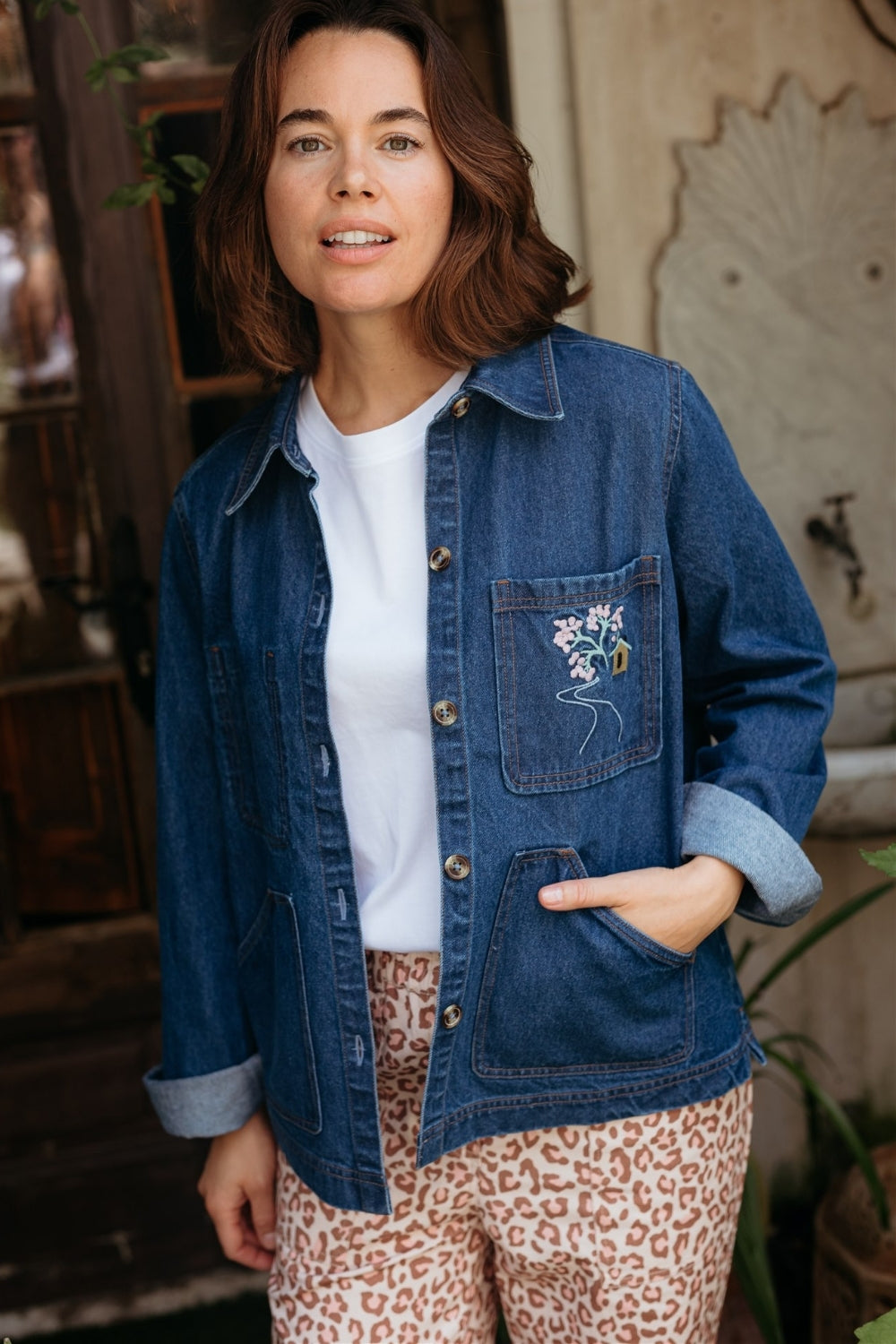 Woman wearing a blue denim jacket with floral pocket detail, white shirt, and patterned pants indoors.