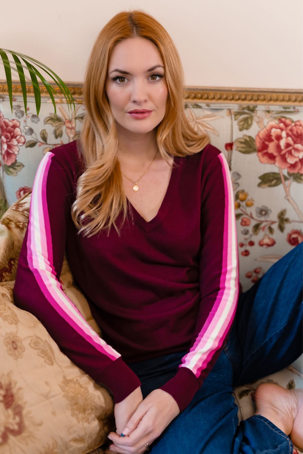 A woman with long blonde hair stands outdoors against a brick wall wearing a long-sleeved midi dress in deep burgundy. The dress has a gathered waist and a small white brand tag near the hem.