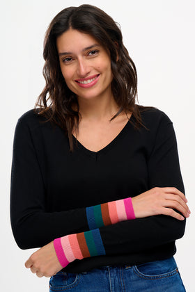 Sugarhill Brighton model wearing a black V-neck jumper with multicoloured stripe cuffs, paired with blue jeans.