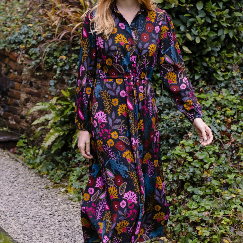 A woman wearing a long-sleeved midi dress with a colorful floral and botanical print in pink, orange, yellow, and blue on a dark background, standing on a gravel path surrounded by green garden foliage.