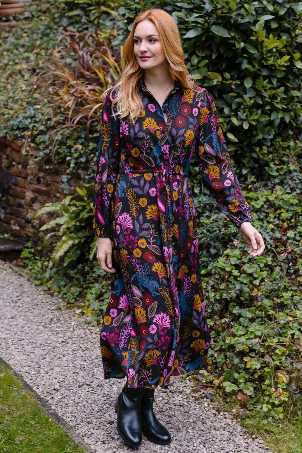 A woman wearing a long-sleeved midi dress with a colorful floral and botanical print in pink, orange, yellow, and blue on a dark background, standing on a gravel path surrounded by green garden foliage.