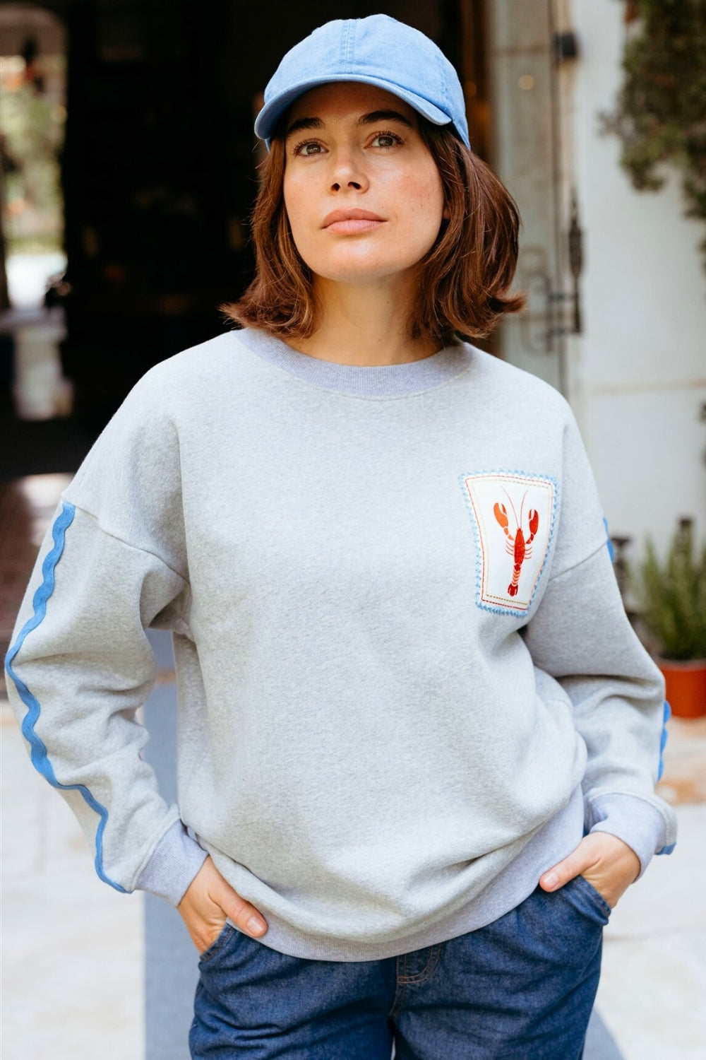 Person wearing a light gray sweatshirt with a patch and a blue cap outdoors.