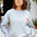 Person wearing a light gray sweatshirt with a patch and a blue cap outdoors.