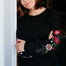 Woman wearing a black sweatshirt with floral designs and teal pants on a white background
