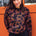 A woman wearing a black sweatshirt with a two-tone leopard print pattern, paired with black trousers, standing in front of an orange wall.