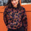A woman wearing a black sweatshirt with a two-tone leopard print pattern, paired with black trousers, standing in front of an orange wall.