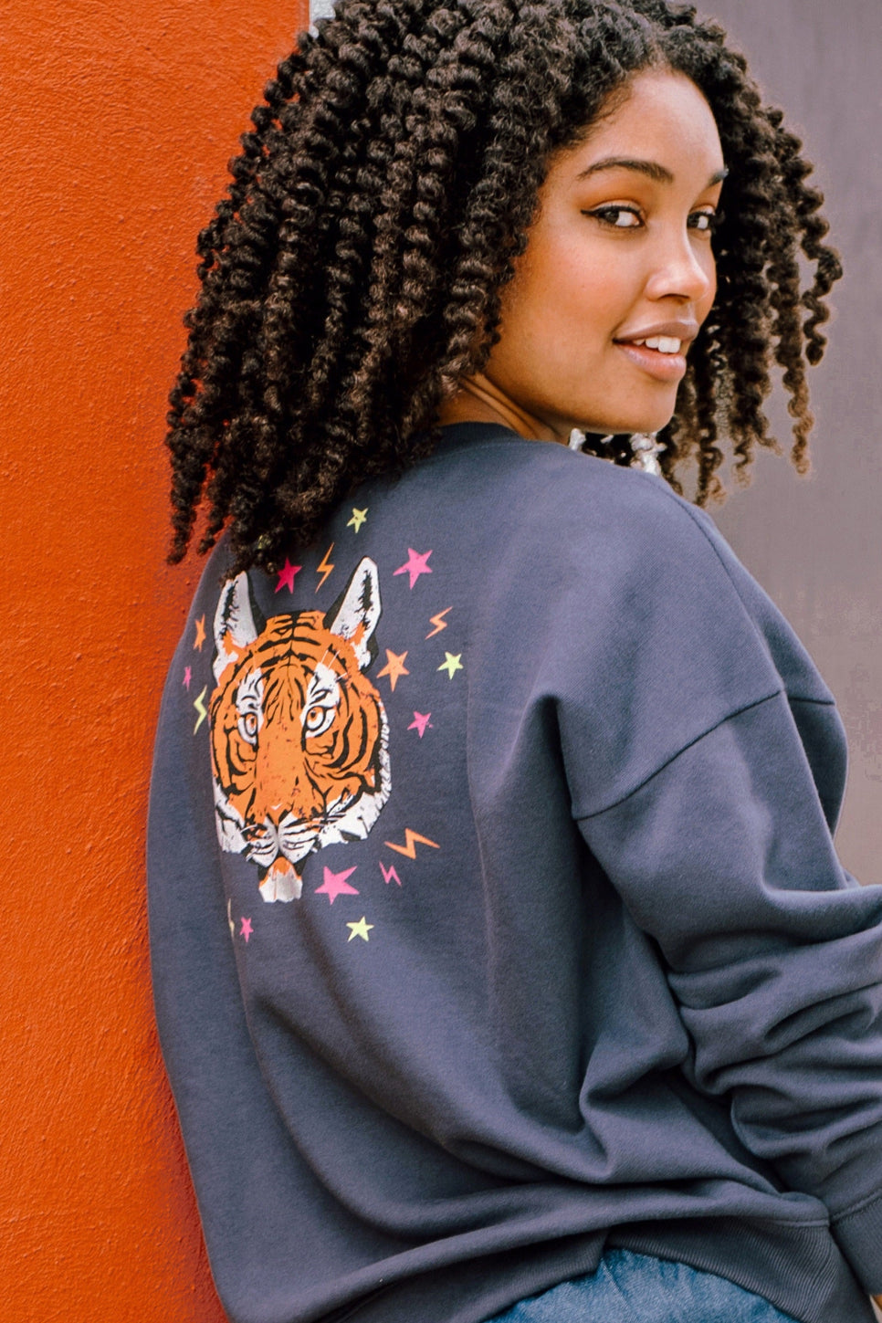 A woman wearing an oversized sweatshirt with a crew neck, featuring a printed tiger face with stars and lightning bolts in pink, neon green, and orange on a charcoal blue base.