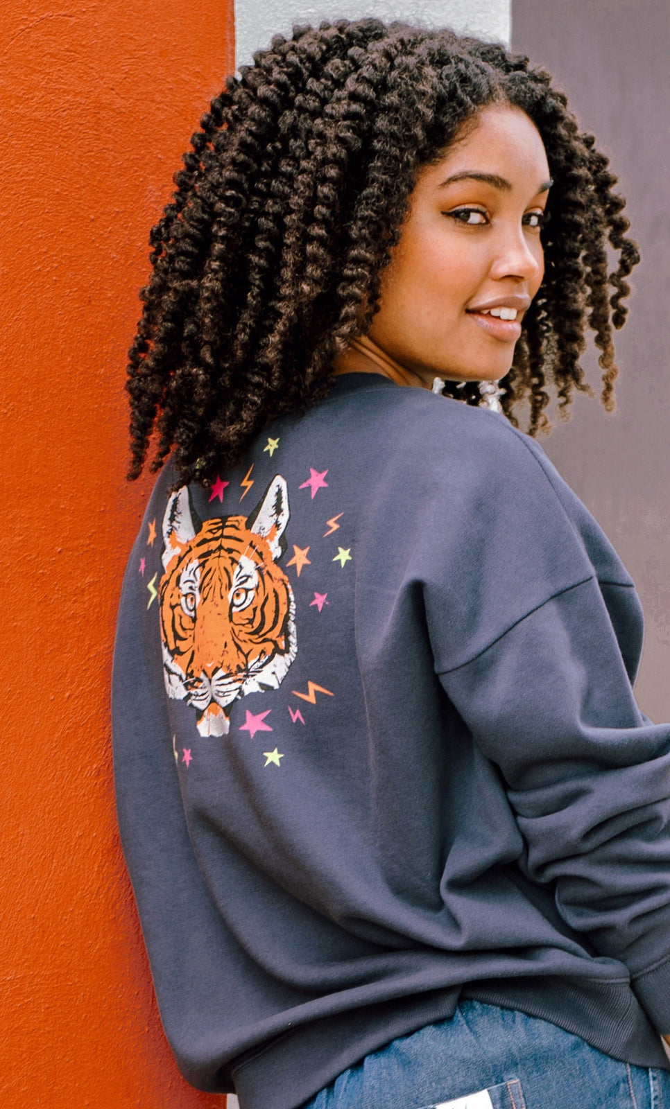 A woman wearing an oversized sweatshirt with a crew neck, featuring a printed tiger face with stars and lightning bolts in pink, neon green, and orange on a charcoal blue base.