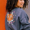 A woman wearing an oversized sweatshirt with a crew neck, featuring a printed tiger face with stars and lightning bolts in pink, neon green, and orange on a charcoal blue base.