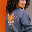 A woman wearing an oversized sweatshirt with a crew neck, featuring a printed tiger face with stars and lightning bolts in pink, neon green, and orange on a charcoal blue base.