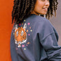 A woman wearing an oversized sweatshirt with a crew neck, featuring a printed tiger face with stars and lightning bolts in pink, neon green, and orange on a charcoal blue base.