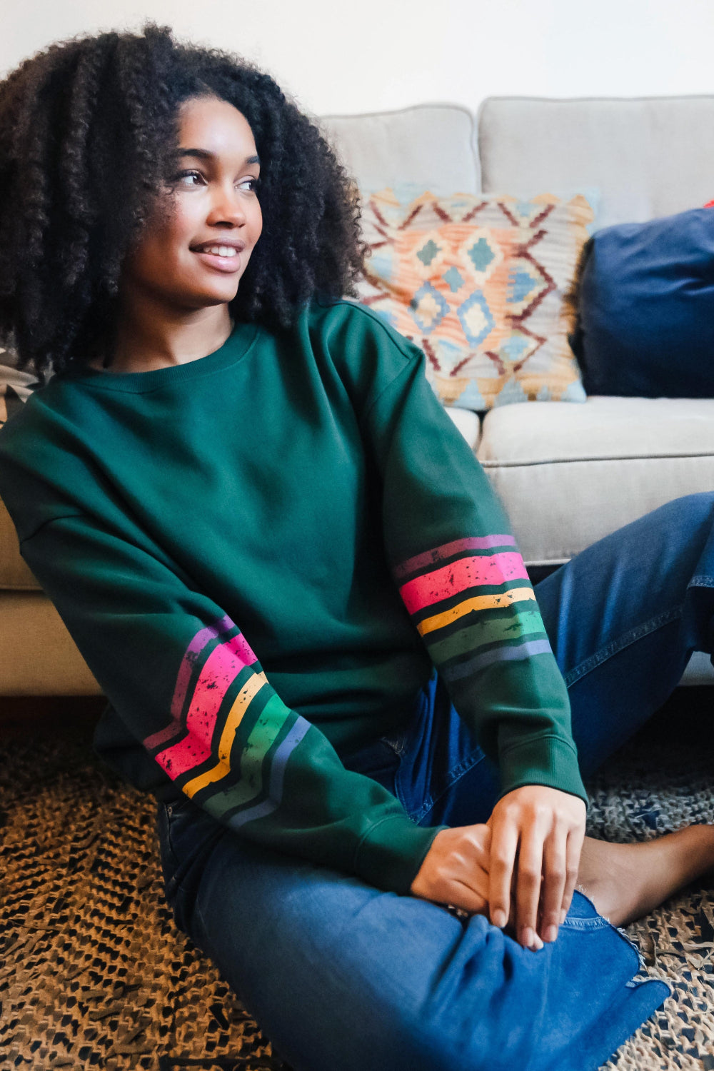 A Sugarhill Brighton model sitting on a sofa wearing a dark green crew neck sweatshirt with colourful striped sleeves.