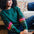 A Sugarhill Brighton model sitting on a sofa wearing a dark green crew neck sweatshirt with colourful striped sleeves.