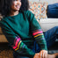 A Sugarhill Brighton model sitting on a sofa wearing a dark green crew neck sweatshirt with colourful striped sleeves.