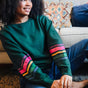 A Sugarhill Brighton model sitting on a sofa wearing a dark green crew neck sweatshirt with colourful striped sleeves.