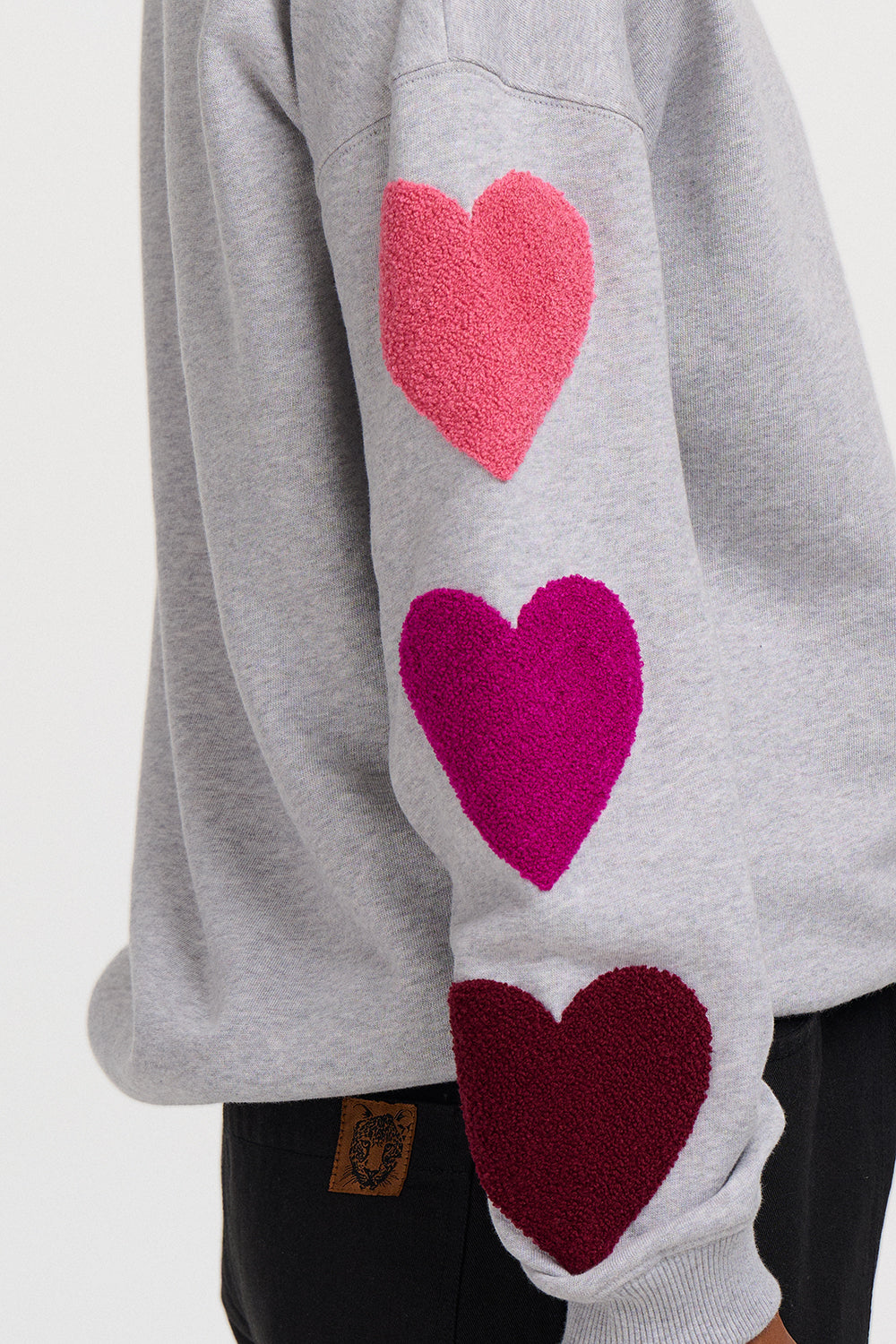 Close-up of a light gray sweatshirt sleeve featuring three large textured heart patches in pink, magenta, and burgundy, worn with black pants.