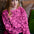 A woman with red hair sits on a brown leather couch wearing a bright pink long-sleeve sweatshirt with a dark pink heart pattern, paired with dark blue pants.