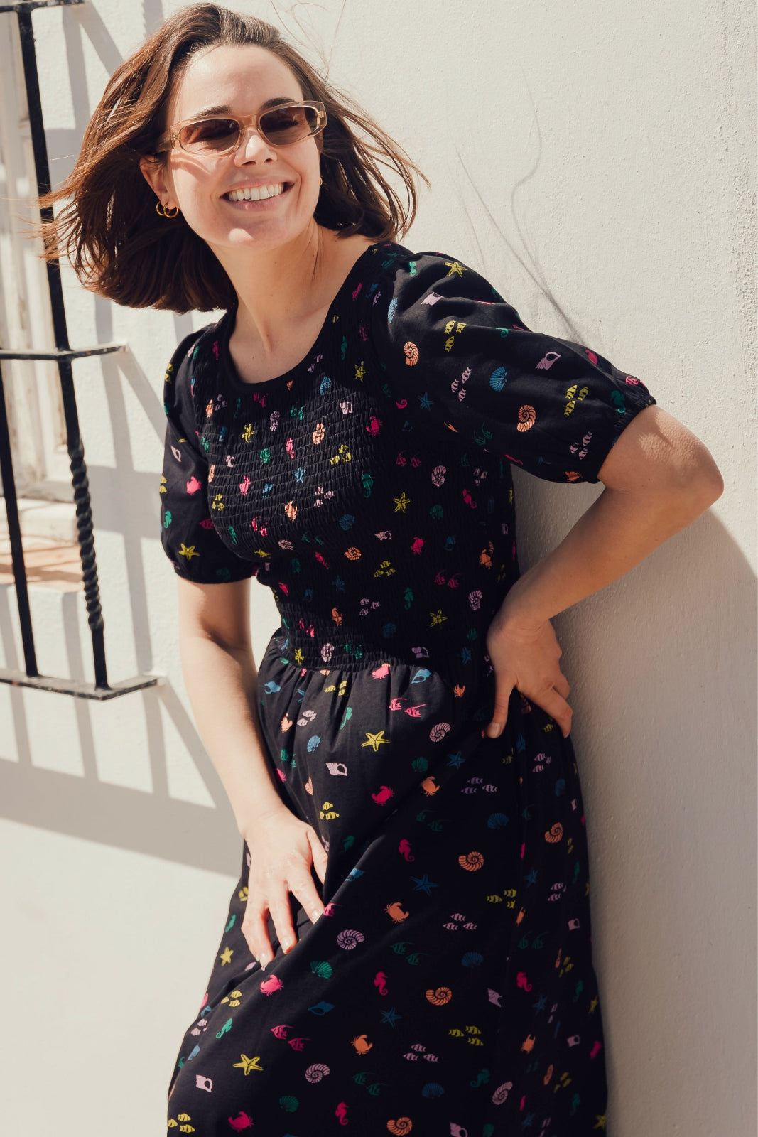 A model wearing a short-sleeved black midi dress with a multicolored underwater theme, including shells and fish, standing outdoors.