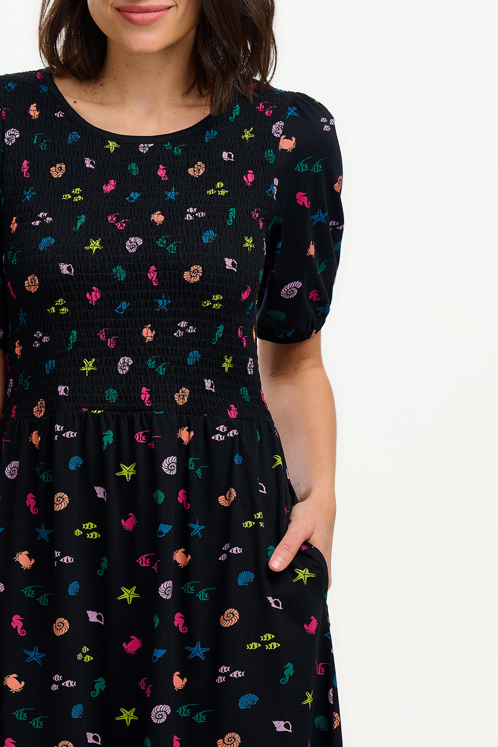 A model wearing a vintage-inspired, black midi dress with a multicolored underwater theme, including small shells and fish on a contrasting black base. The dress features short puff sleeves and a full mini skirt.