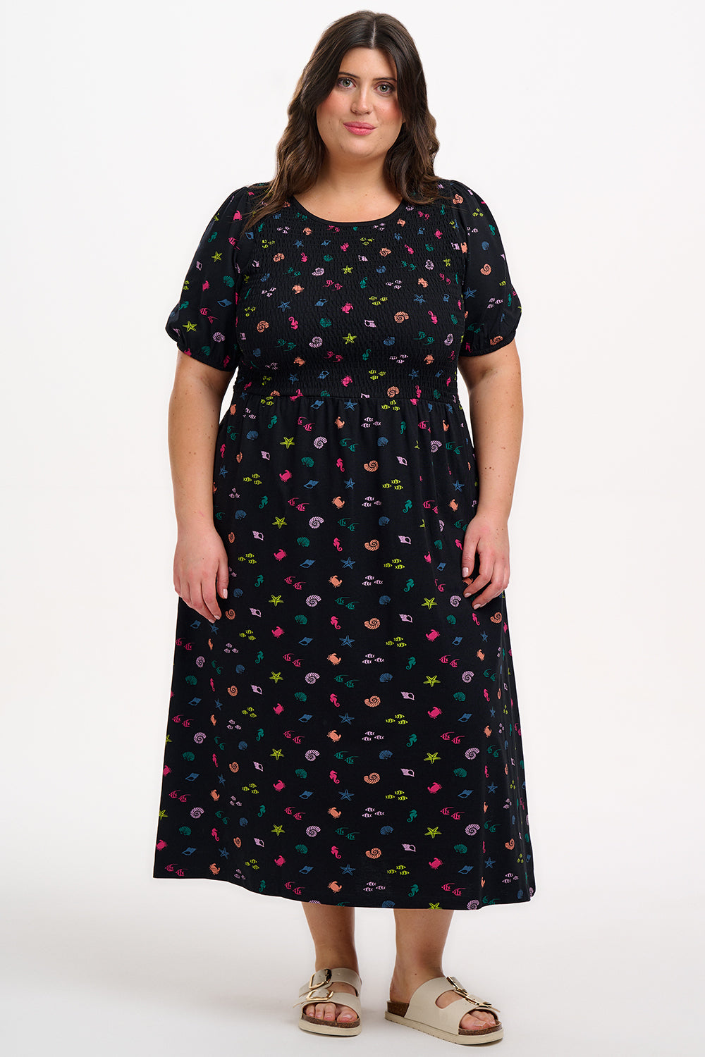 A model wearing a vintage-inspired, black midi dress with a multicolored underwater theme, including small shells and fish on a contrasting black base. The dress features short puff sleeves and a full mini skirt.