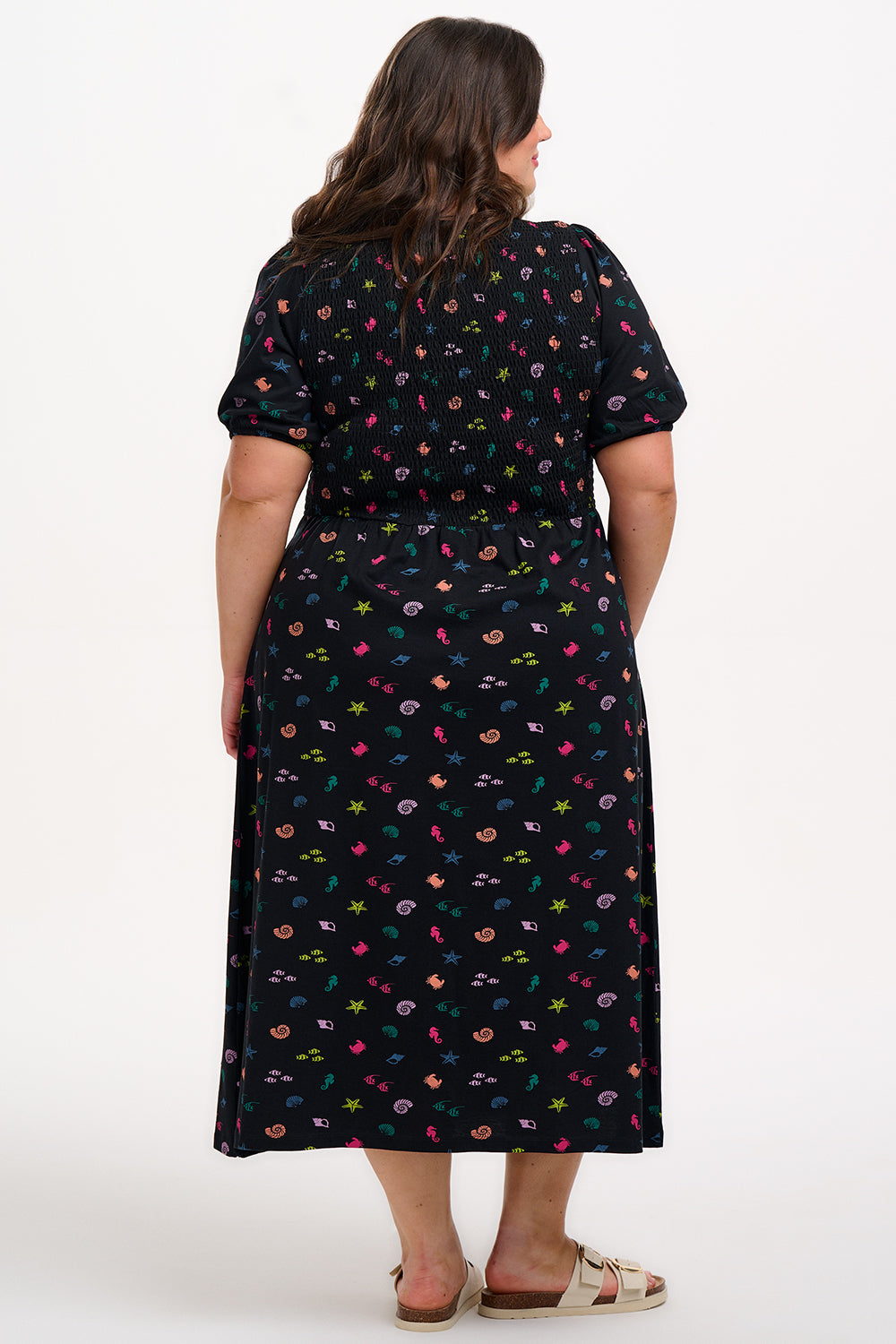 A model wearing a vintage-inspired, black midi dress with a multicolored underwater theme, including small shells and fish on a contrasting black base. The dress features short puff sleeves and a full mini skirt.