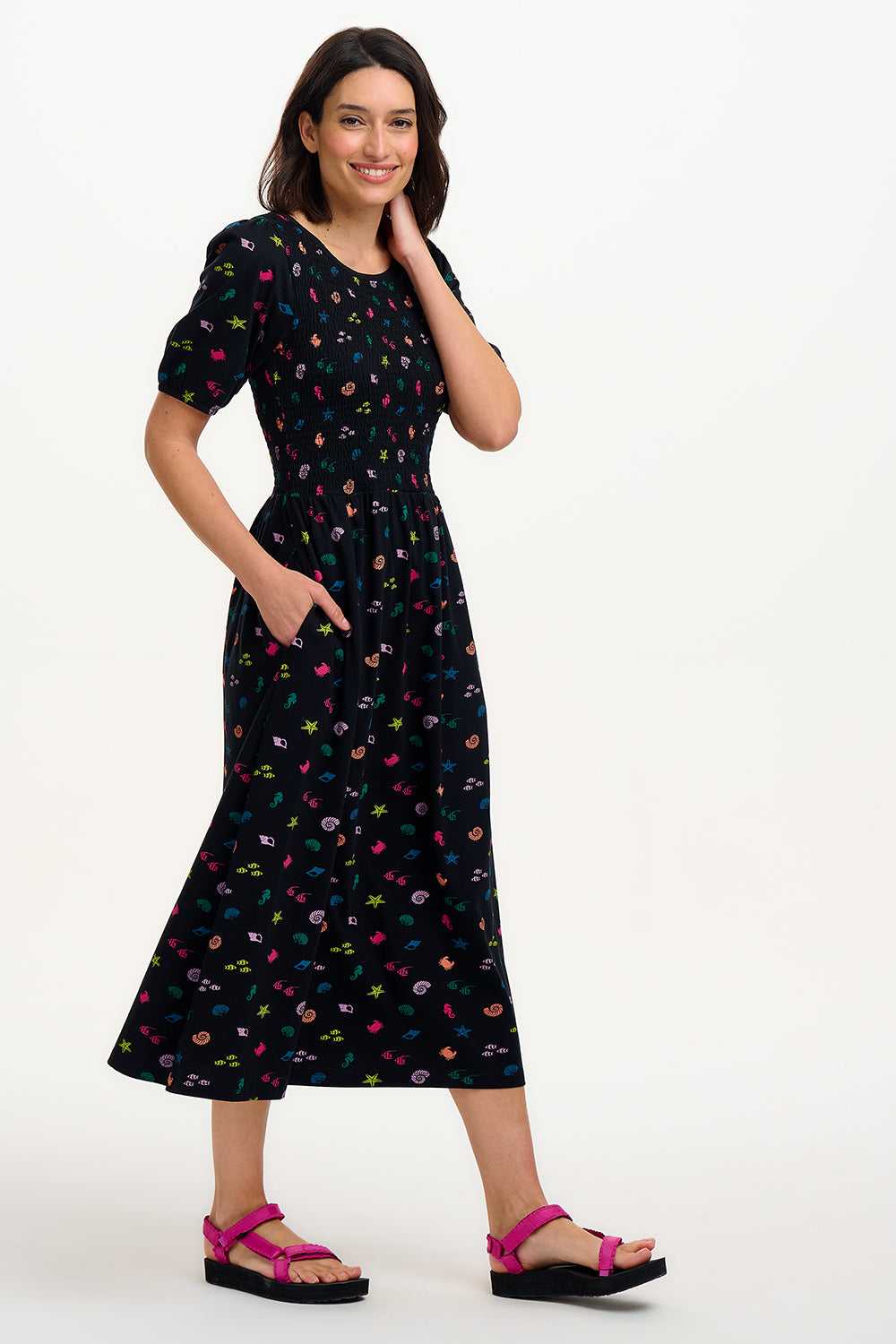 A model wearing a vintage-inspired, black midi dress with a multicolored underwater theme, including small shells and fish on a contrasting black base. The dress features short puff sleeves and a full mini skirt.