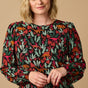 A woman modeling a long-sleeved blouse with a 'Lost Dinosaur' print featuring scenes of dinosaurs among foliage in dark green, sage, tan, mustard, and deep magenta pink colors.