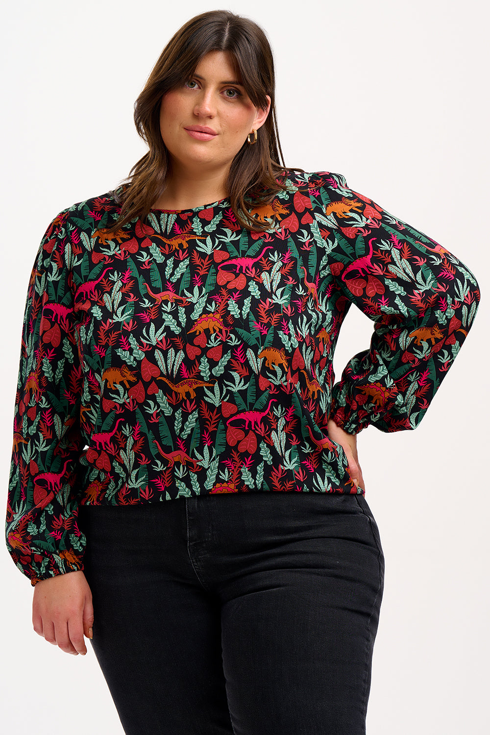 A model wearing a long-sleeved blouse with a round neckline and a keyhole back fastening, featuring a colorful 'Lost Dinosaur' print with scenes of dinosaurs among foliage.