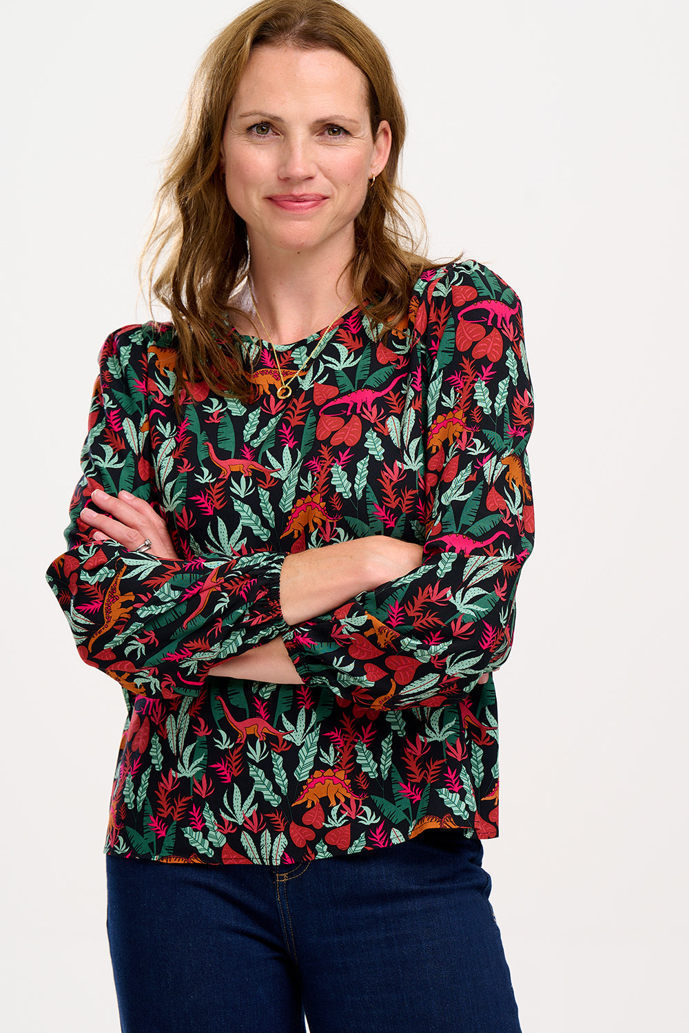 A model wearing a long-sleeved blouse with a round neckline and a keyhole back fastening, featuring a colorful 'Lost Dinosaur' print with scenes of dinosaurs among foliage.