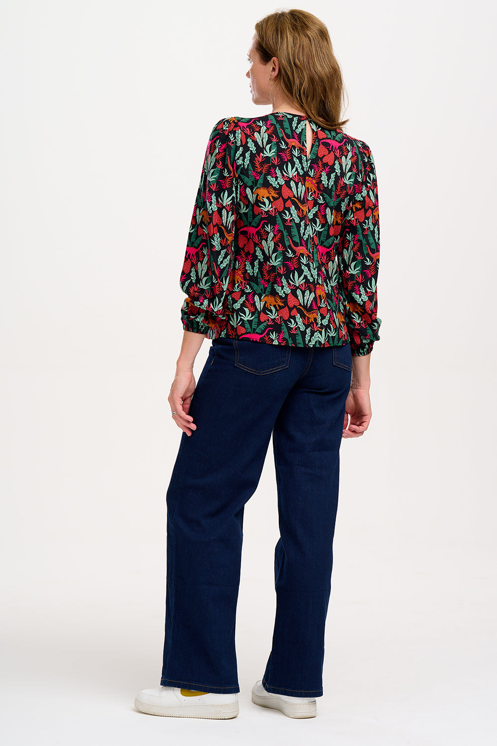 A model wearing a long-sleeved blouse with a round neckline and a keyhole back fastening, featuring a colorful 'Lost Dinosaur' print with scenes of dinosaurs among foliage.