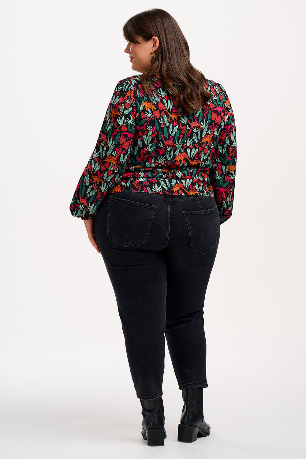 A model wearing a long-sleeved blouse with a round neckline and a keyhole back fastening, featuring a colorful 'Lost Dinosaur' print with scenes of dinosaurs among foliage.