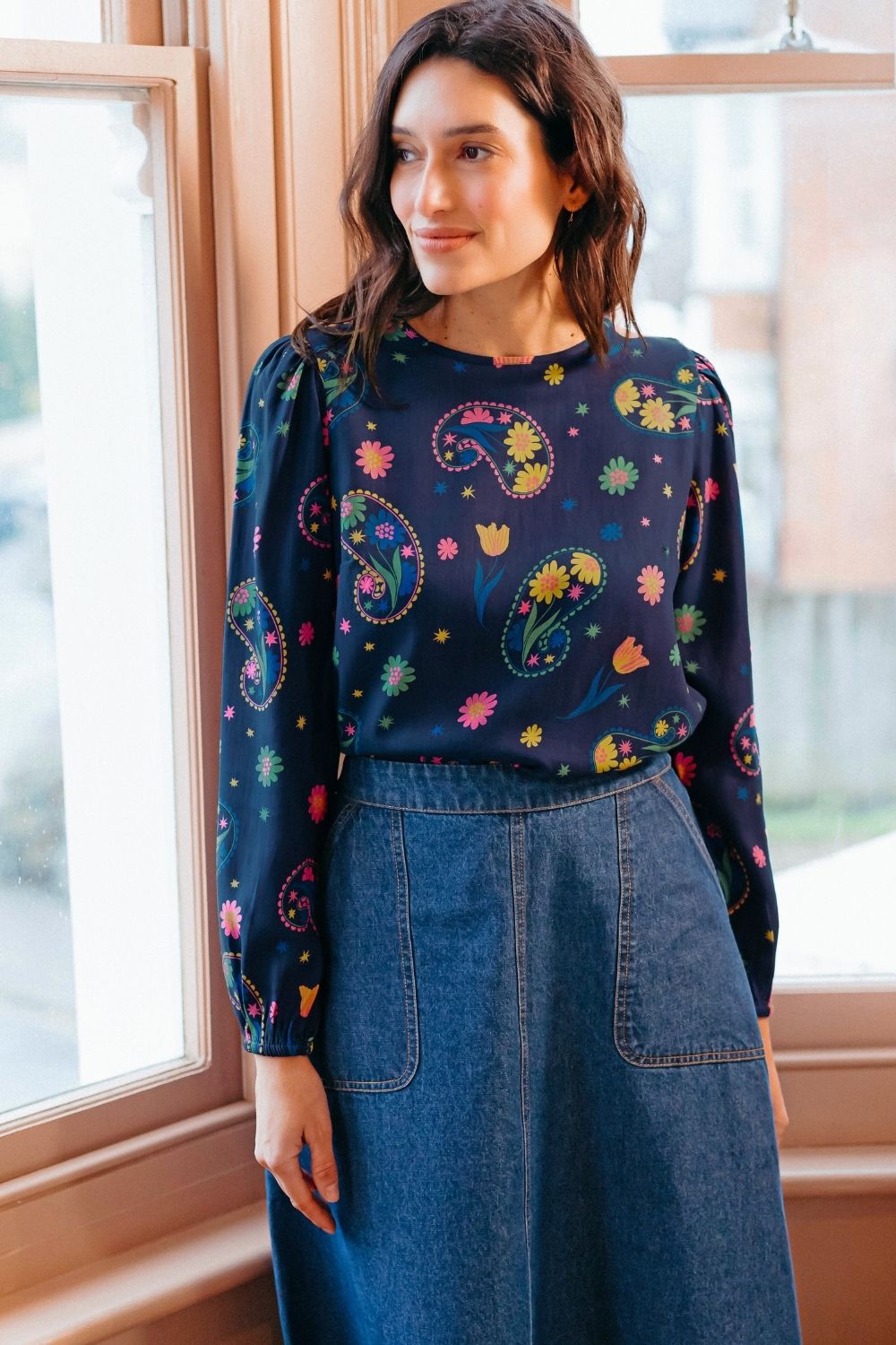Sugarhill Brighton model wearing a colourful patterned top and blue denim skirt indoors.