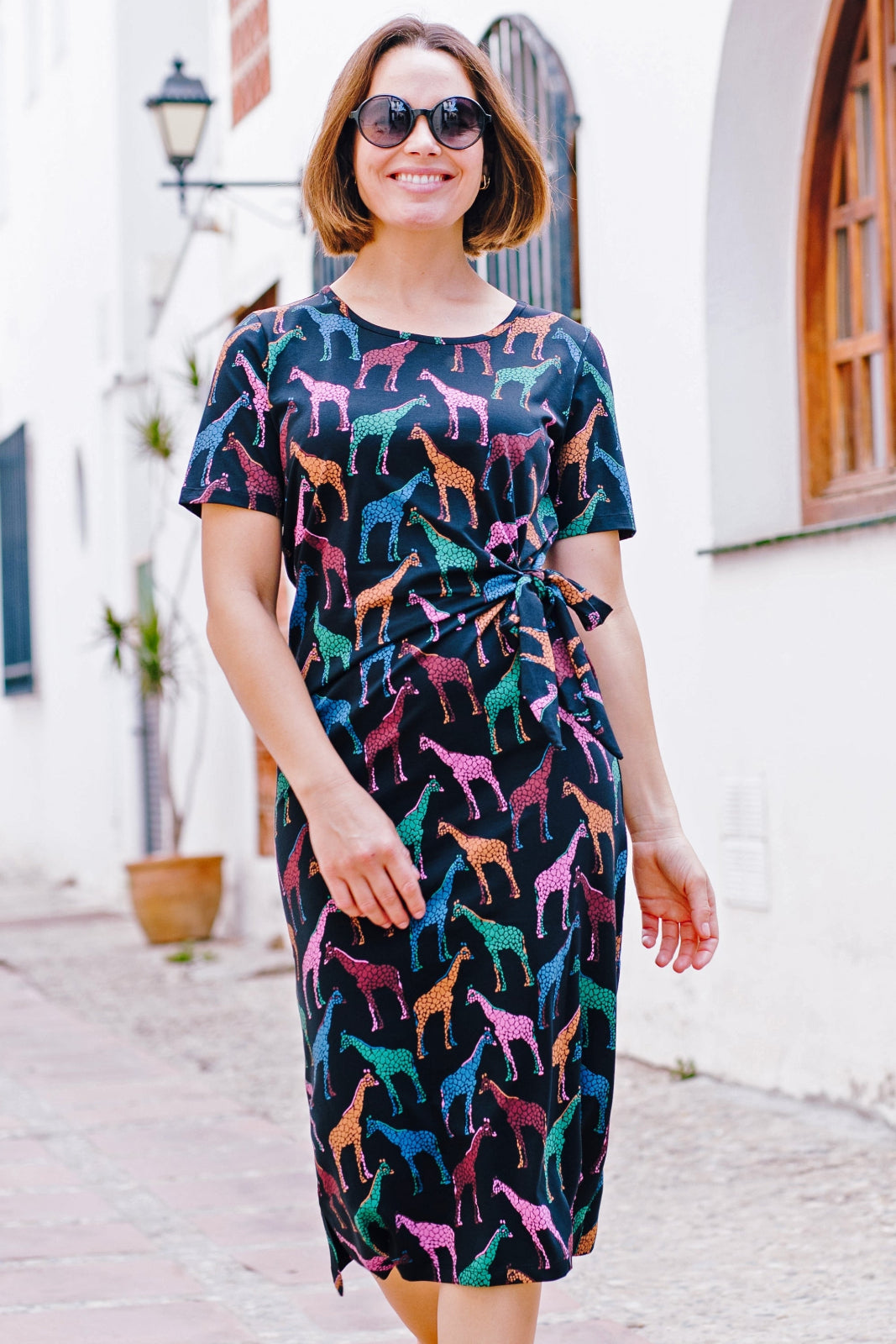A woman standing on a street wearing a midi jersey dress with a giraffe print in multiple colors on a black base.
