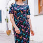 A woman standing on a street wearing a midi jersey dress with a giraffe print in multiple colors on a black base.