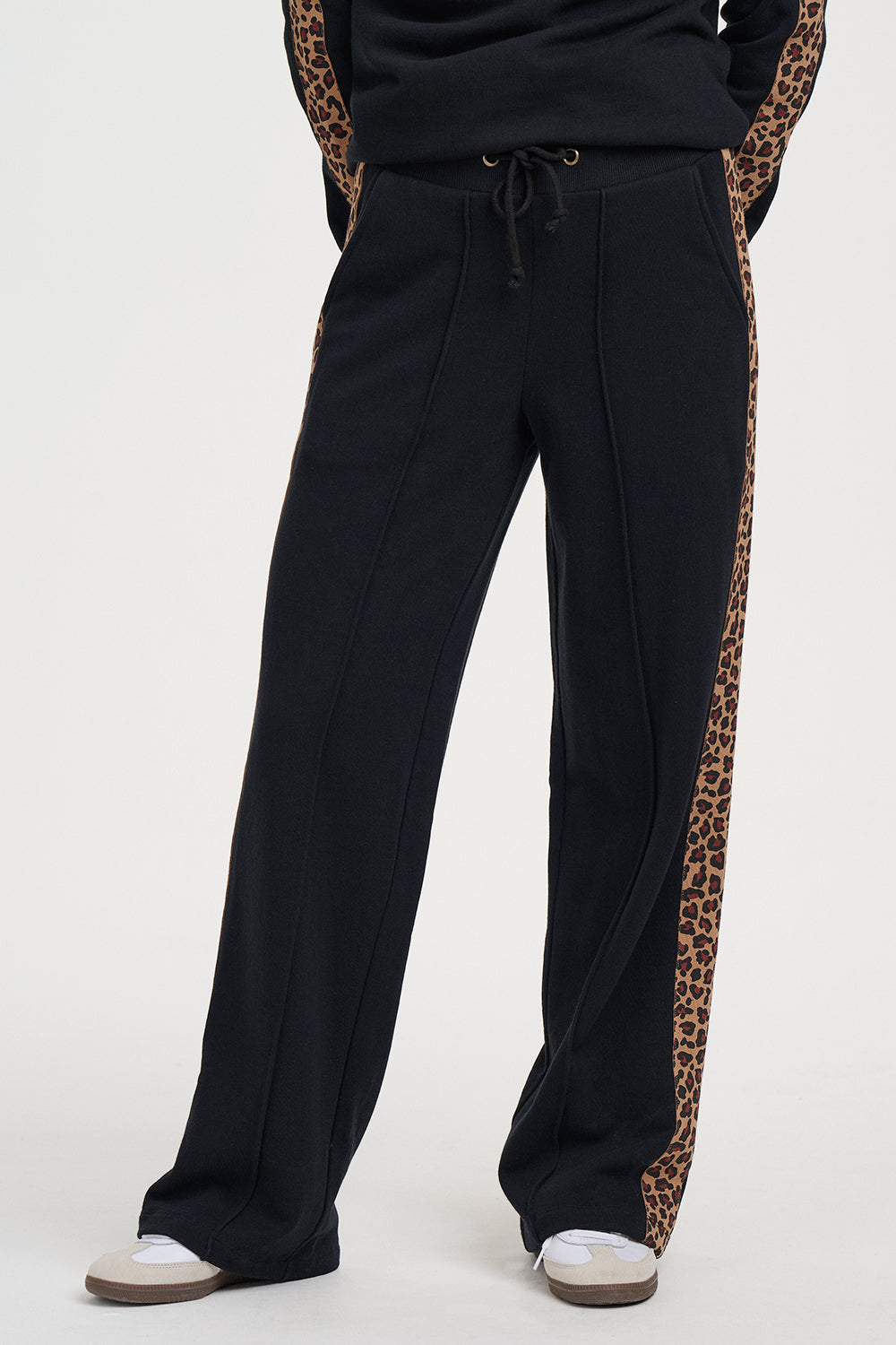 Black wide-leg sweatpants with leopard print side panels, shown on a model from the waist down wearing white sneakers against a light gray studio background.