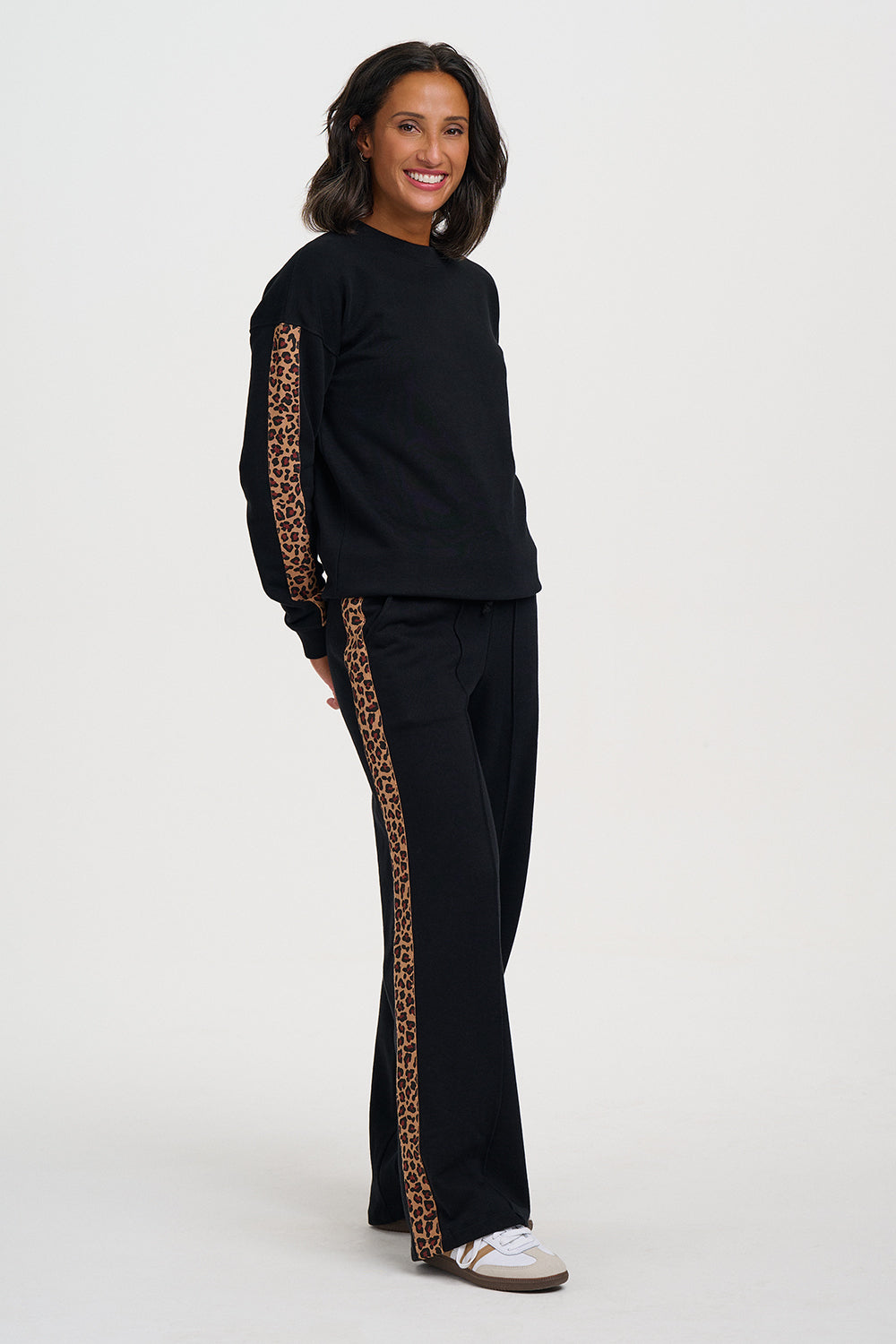 A woman modeling a black two-piece lounge set with leopard print side panels, consisting of a long-sleeve top and matching wide-leg pants, paired with white sneakers against a plain white studio background.