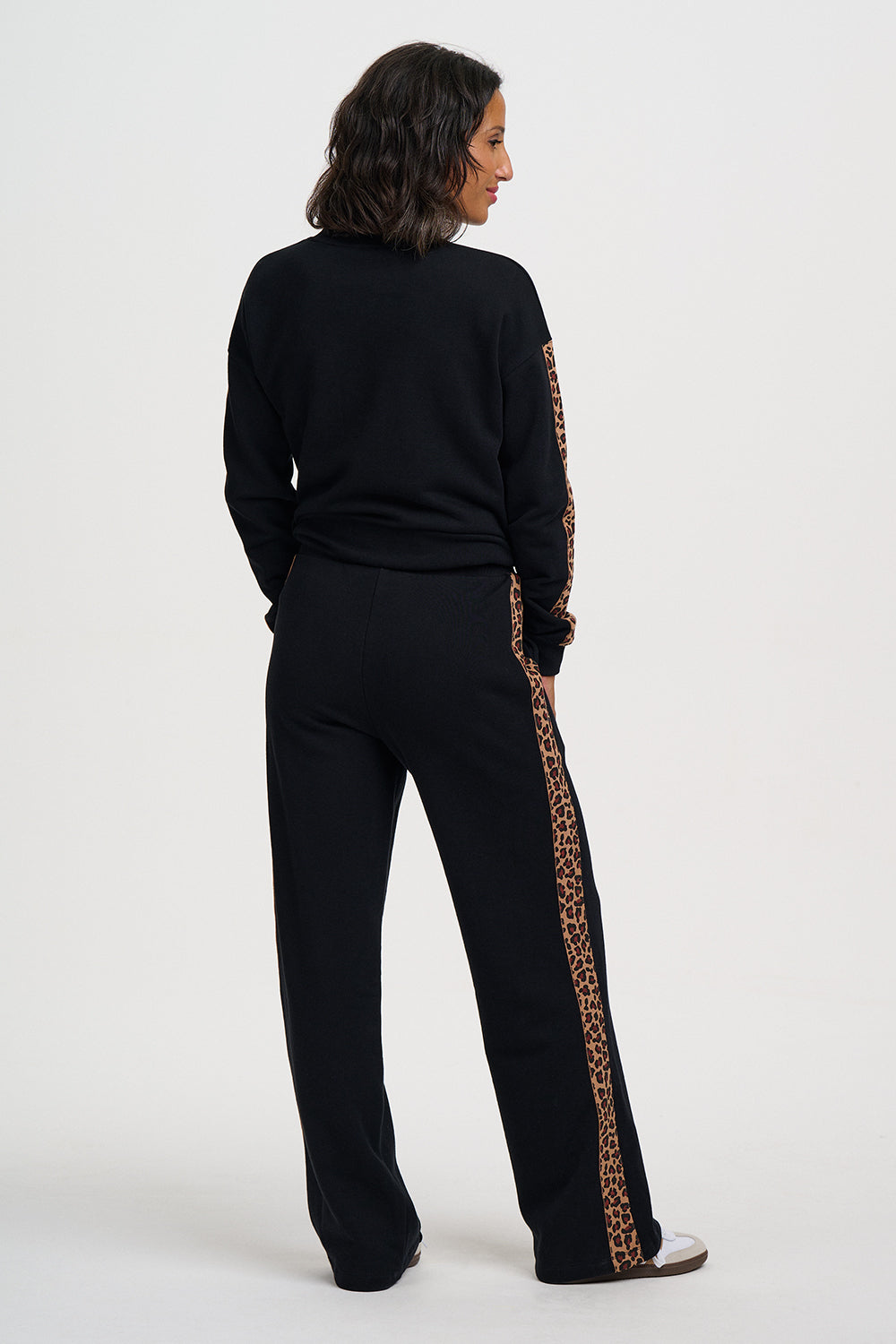 A woman shown from the back wearing a black knit tracksuit with leopard-print side stripes, photographed against a plain light gray studio background.