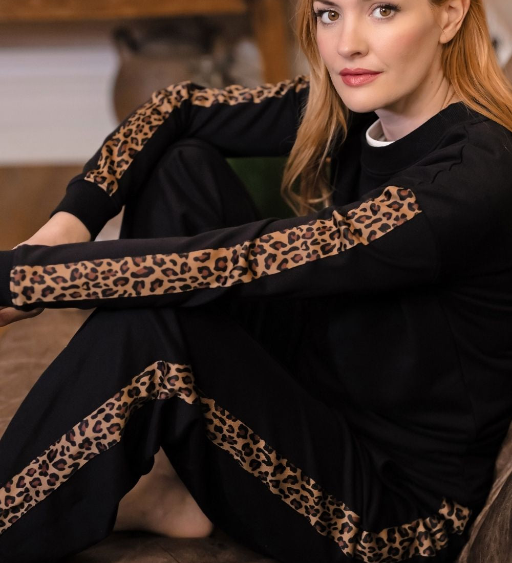A woman with red hair sits on a brown couch wearing a black long-sleeve pajama set with leopard print stripes on the sleeves and legs.