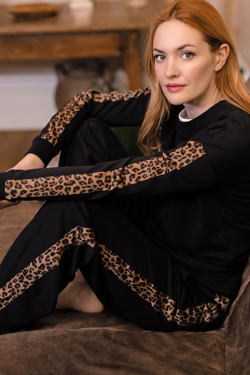 A woman with red hair sits on a brown couch wearing a black long-sleeve pajama set with leopard print stripes on the sleeves and legs.