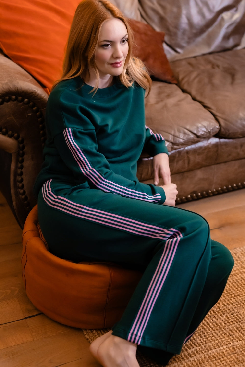 A woman with red hair sits barefoot on an orange pouf in a living room, wearing a dark teal green sweatshirt and matching wide-leg sweatpants with pink and white stripes down the sleeves and legs.