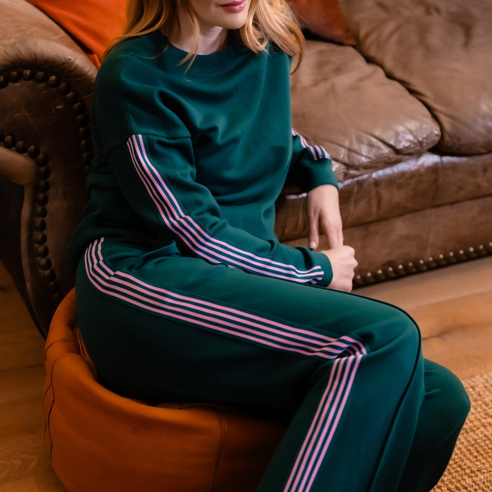 A woman with red hair sits barefoot on an orange pouf in a living room, wearing a dark teal green sweatshirt and matching wide-leg sweatpants with pink and white stripes down the sleeves and legs.