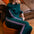 A woman with red hair sits barefoot on an orange pouf in a living room, wearing a dark teal green sweatshirt and matching wide-leg sweatpants with pink and white stripes down the sleeves and legs.