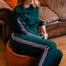 A woman with red hair sits barefoot on an orange pouf in a living room, wearing a dark teal green sweatshirt and matching wide-leg sweatpants with pink and white stripes down the sleeves and legs.