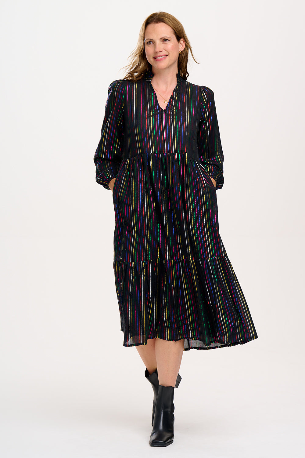 A model wearing a midi smock dress with black metallic rainbow stripes and a V-neck collar, paired with ankle boots.