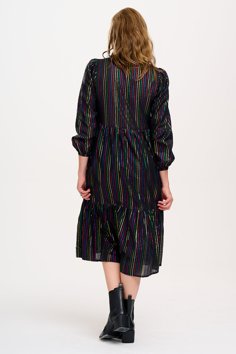A model wearing a midi smock dress with black metallic rainbow stripes and a V-neck collar, paired with ankle boots.