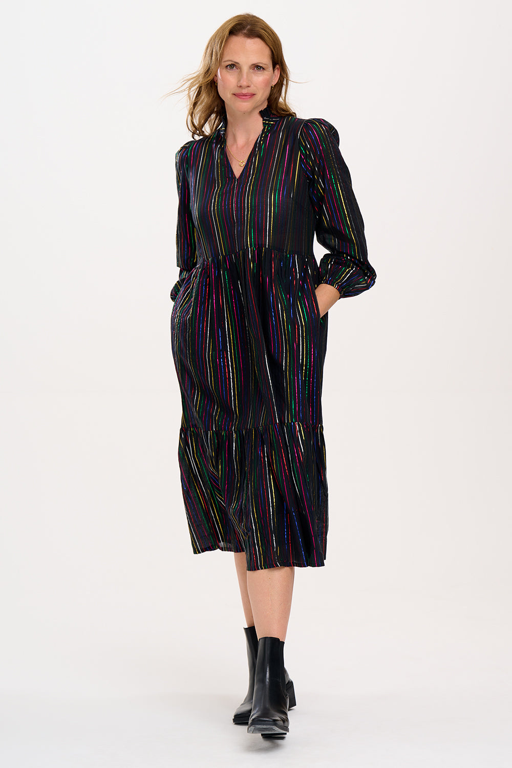 A model wearing a midi smock dress with black metallic rainbow stripes and a V-neck collar, paired with ankle boots.